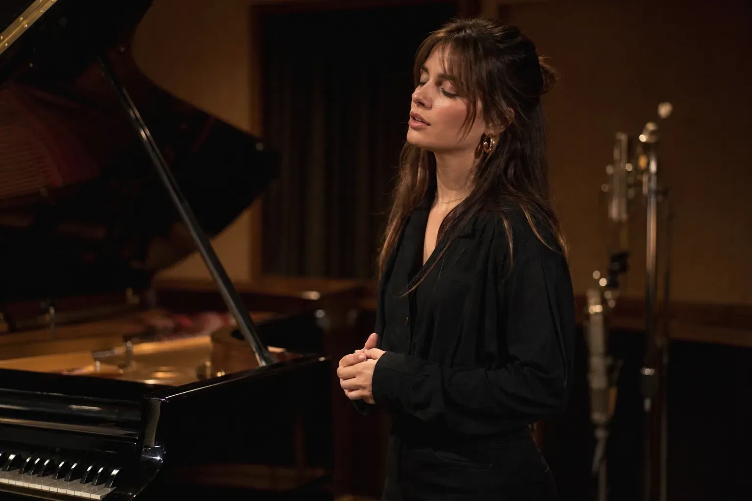Une jeune chanteuse devant un piano dans un studio, éclairée par une lumière douce, dans une ambiance mélancolique et intimiste.