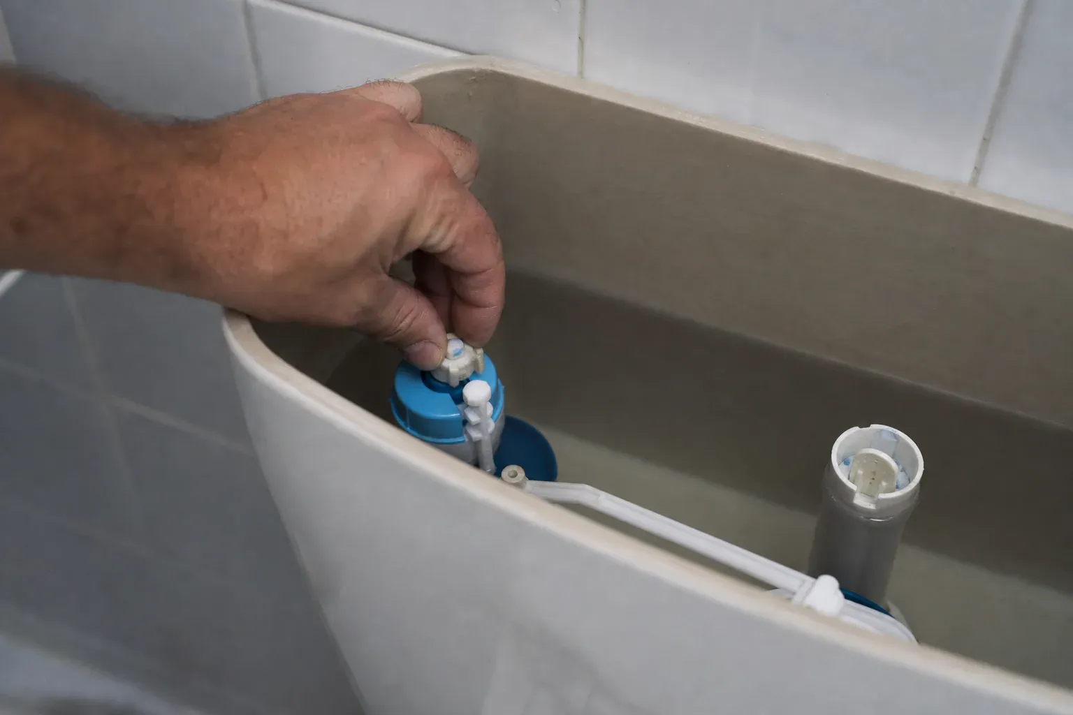Réservoir de chasse d'eau en céramique ouvert, montrant le robinet flotteur et le mécanisme de chasse, une main réglant le flotteur.