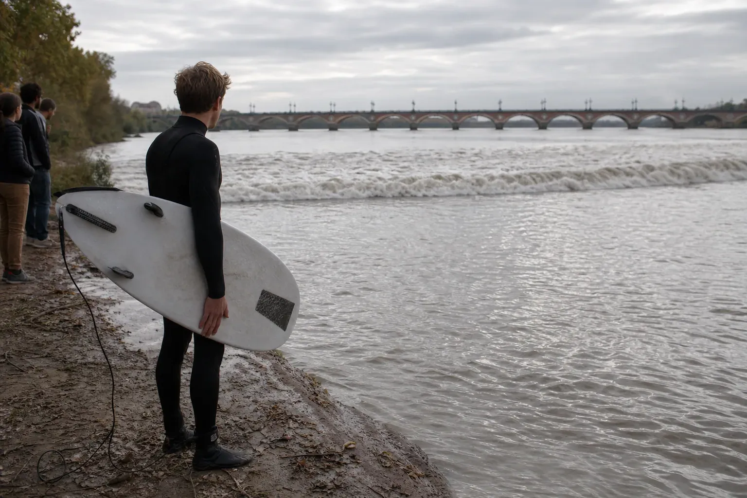 Un surfeur débutant observe le mascaret de la Garonne depuis une berge, planche à la main, avec le fleuve en crue et des spectateurs au loin.
