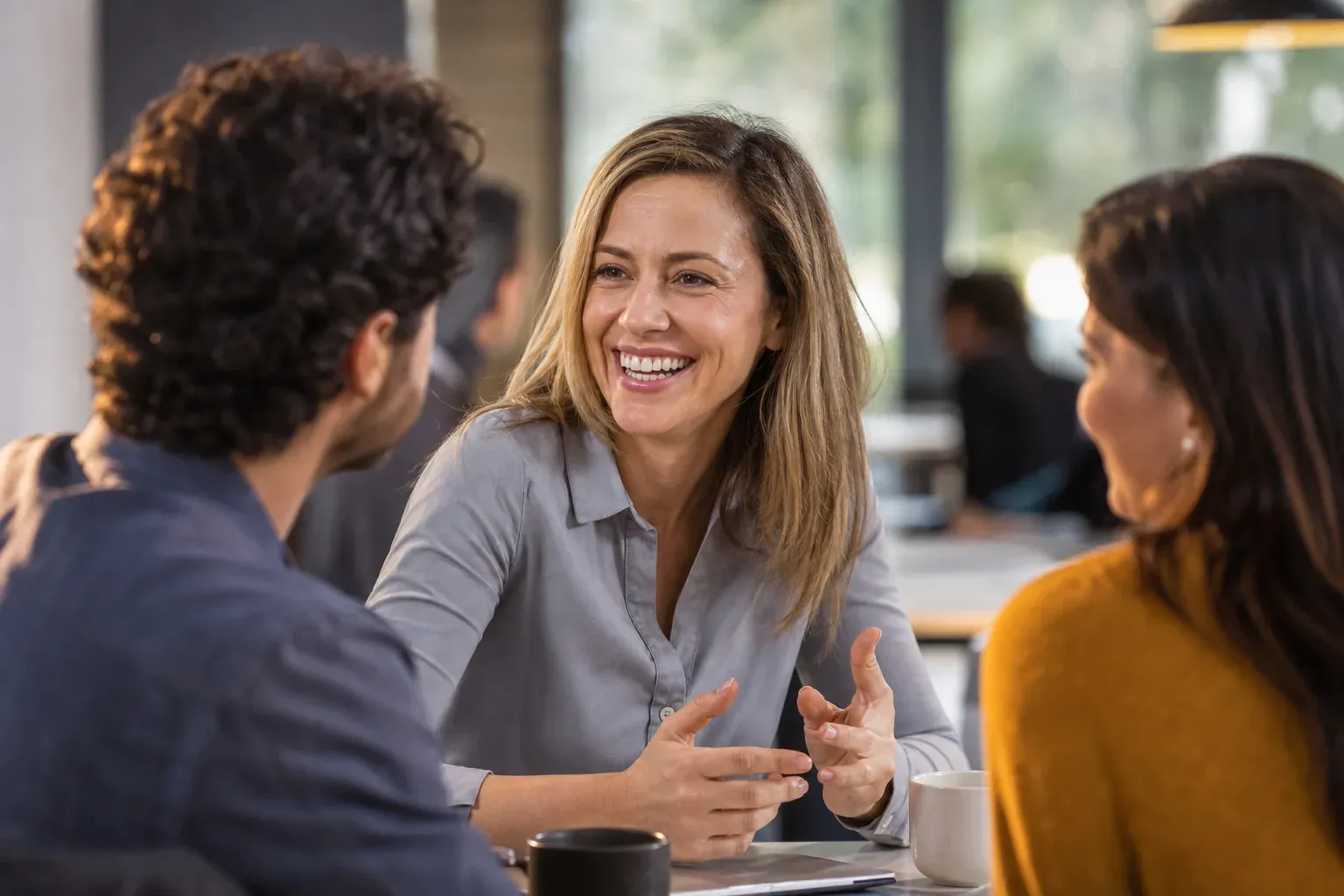 Une personne échange naturellement avec deux interlocuteurs dans un café lumineux, avec une posture ouverte et un sourire sincère.