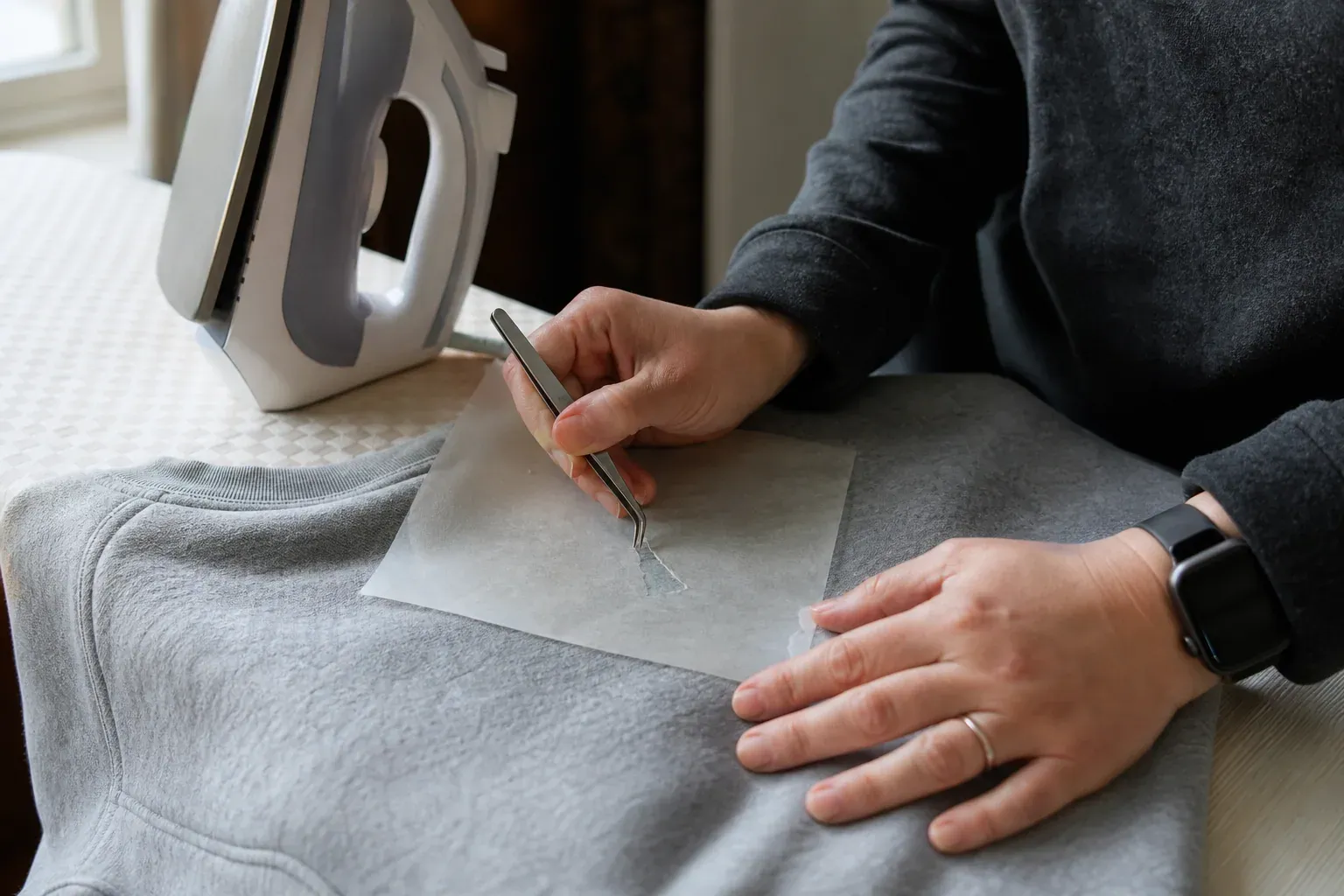 Un vêtement posé sur une planche à repasser avec un motif thermocollé en cours de retrait, du papier cuisson et un fer à repasser à côté.