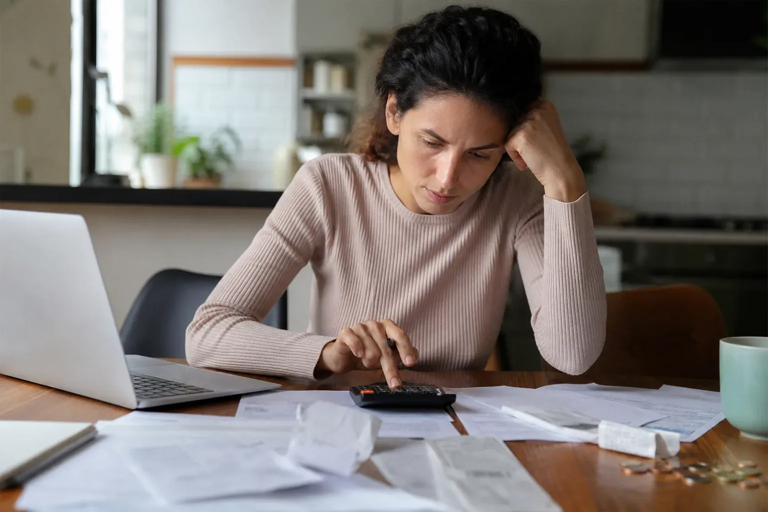 Personne comparant des relevés bancaires et des tickets de caisse sur une table de cuisine, avec calculatrice, ordinateur portable et pièces en euros.