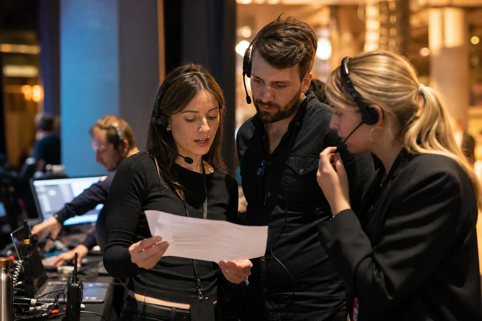 Équipe d'agence événementielle en backstage, autour d'une table de régie, en train de coordonner un événement avec ordinateurs, talkies-walkies et planning papier.