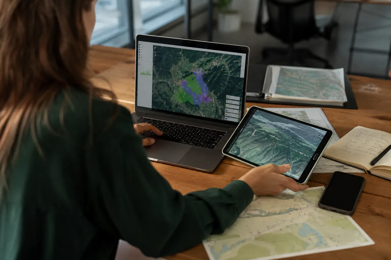 Personne travaillant sur un projet géographique avec une carte sur ordinateur, tablette et documents de terrain sur un bureau.