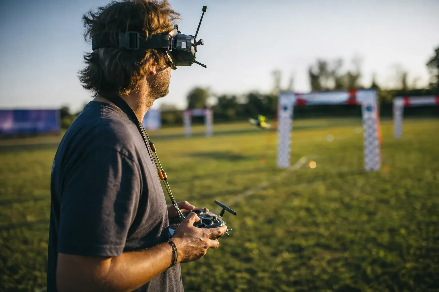 Pilote de drone FPV portant des lunettes immersives et tenant une radiocommande près d’un tracé de course avec des portes en extérieur.