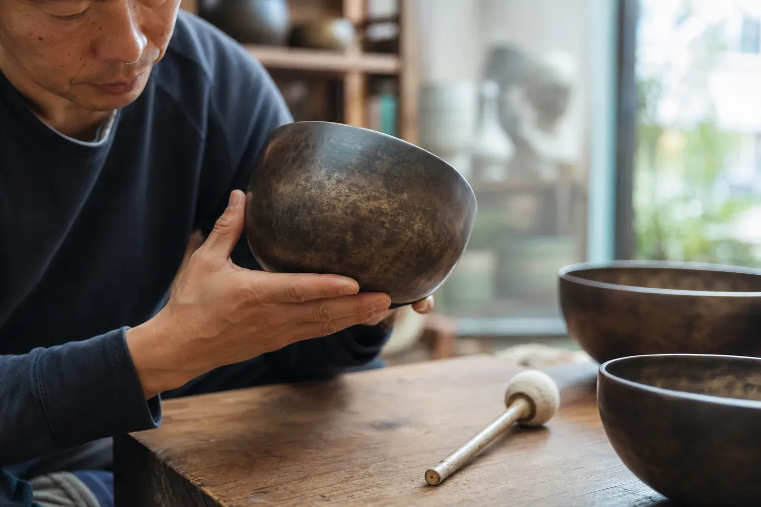 Une personne examine attentivement un bol tibétain martelé à la main sur une table en bois, dans une lumière naturelle douce.