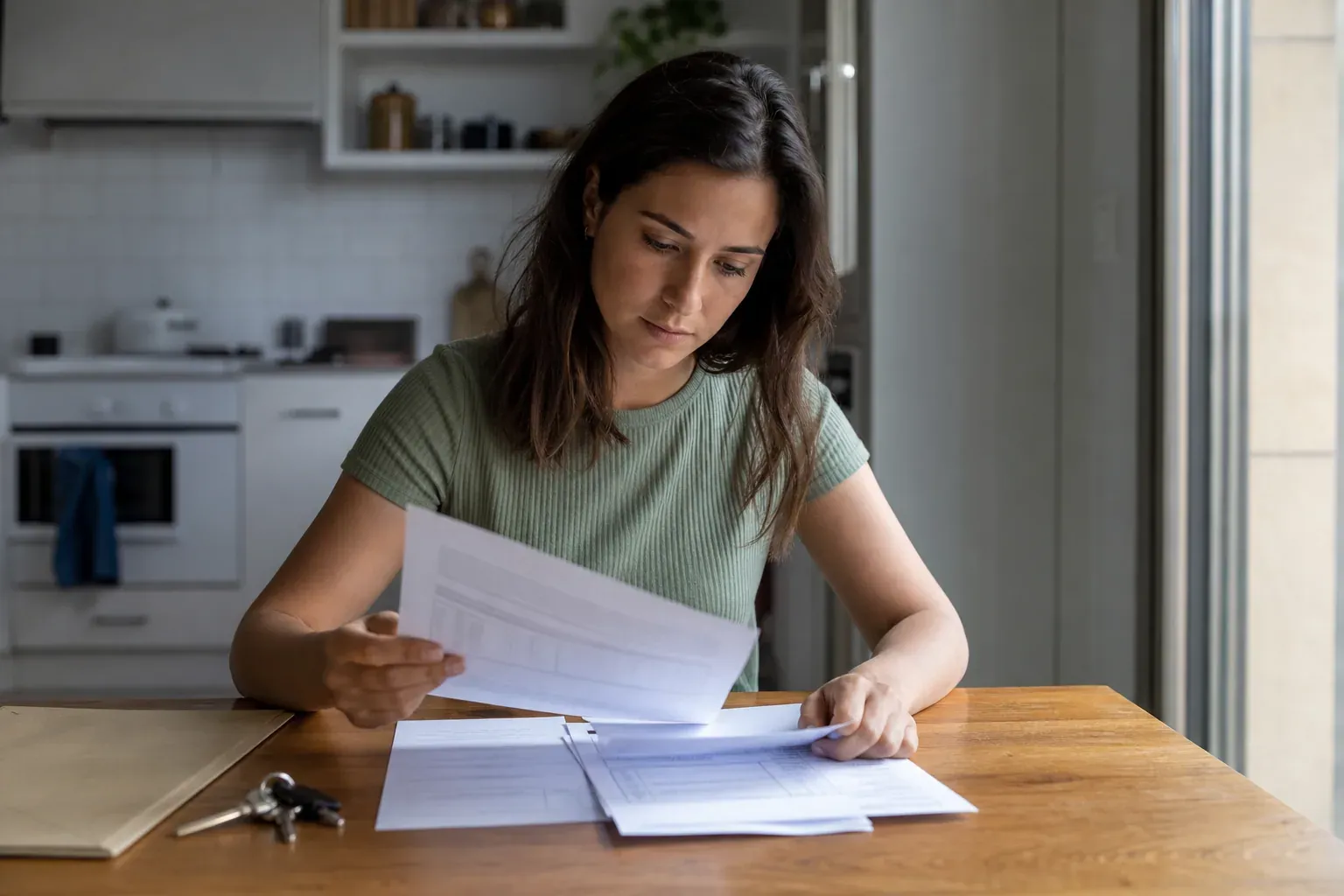 Locataire consultant des documents de bail et un relevé de compte à une table de cuisine, clés d'appartement posées devant lui, lumière naturelle.