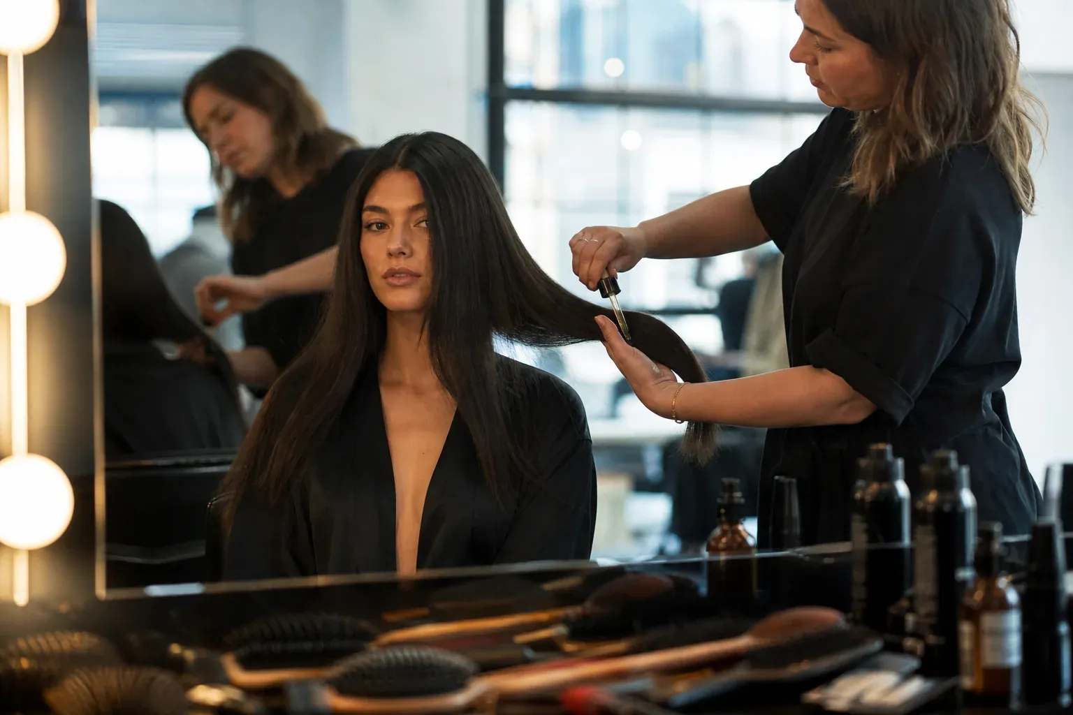 Une femme aux cheveux brillants dans une loge de coiffure, pendant qu’un coiffeur applique un soin naturel, ambiance réaliste et éditoriale.