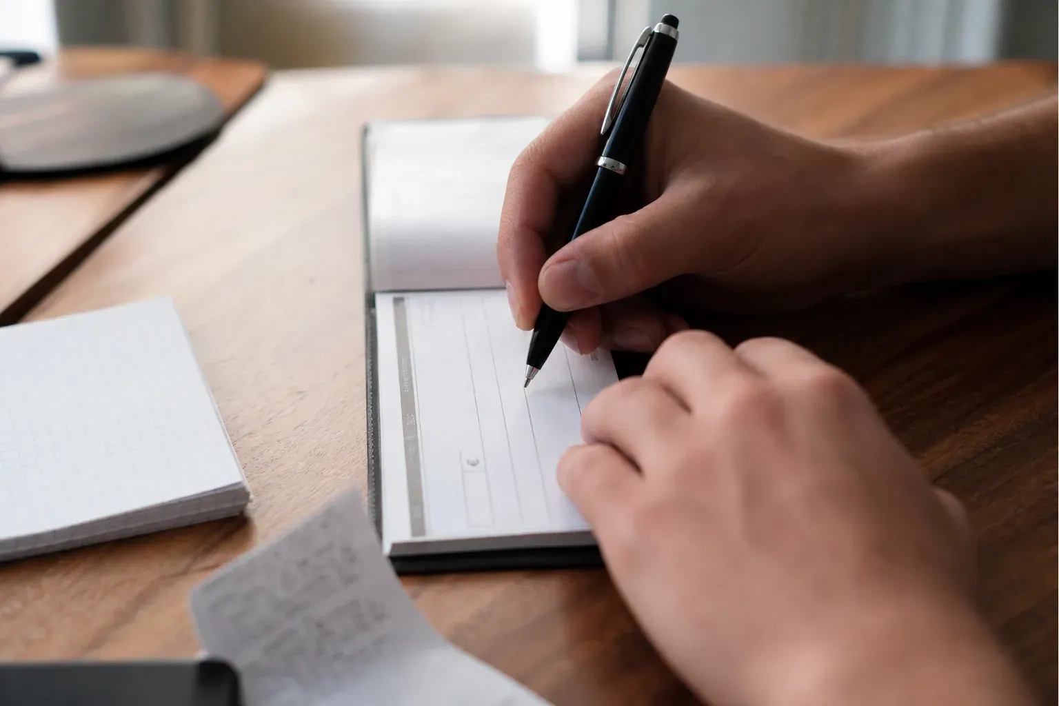 Main en train de remplir un chèque avec un stylo noir, carnet de chèques ouvert sur un bureau, avec des papiers de paiement à côté.