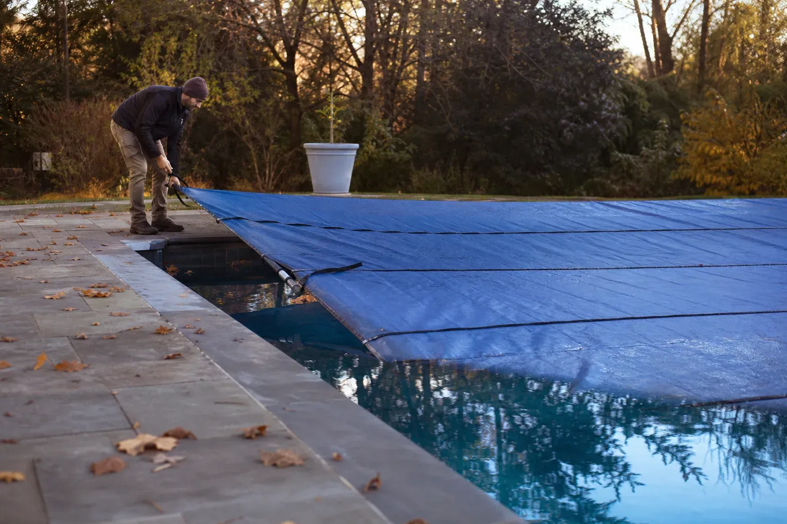 Personne tirant une bâche d'hivernage sur une piscine enterrée au bord de laquelle reposent des feuilles d'automne.