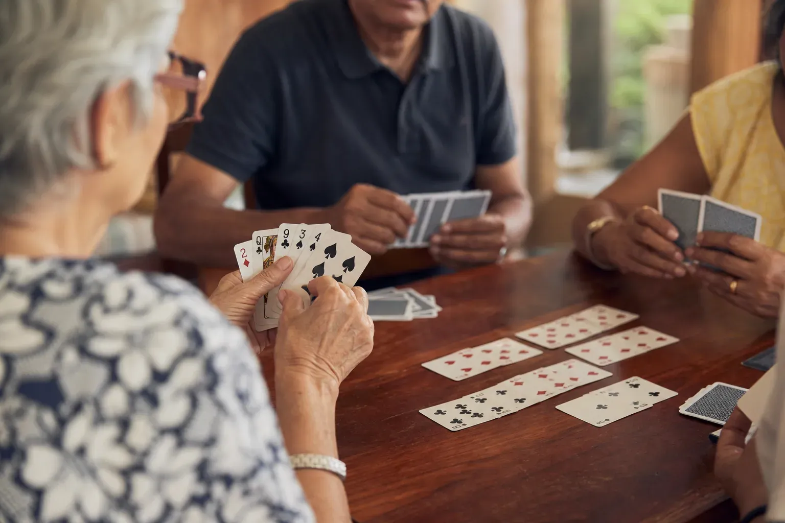 Plusieurs joueurs autour d’une table en bois, avec des cartes de rami tenues en main et des combinaisons posées au centre.