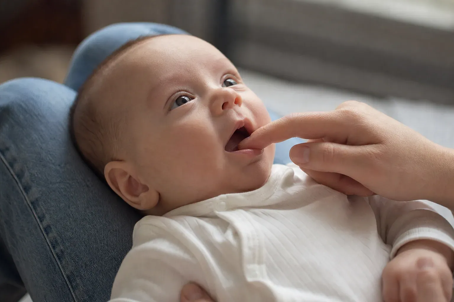 Gros plan d’un bébé de 3 mois allongé dans les bras d’un parent, qui lui masse doucement la bouche dans un intérieur lumineux.