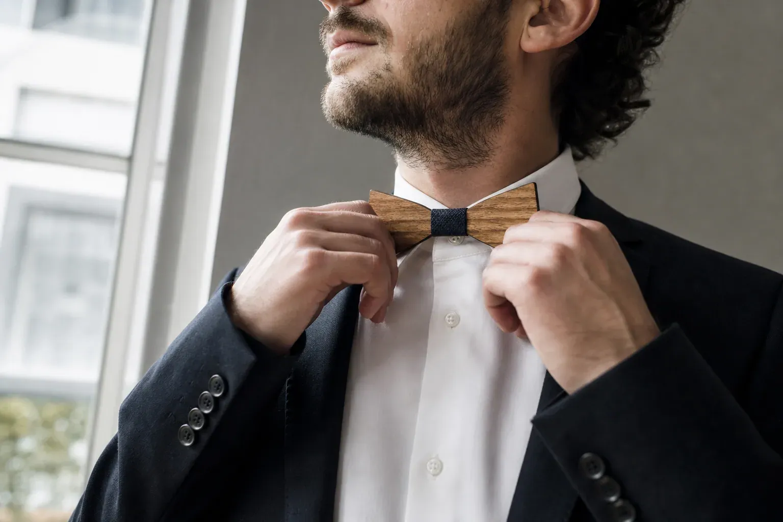 Homme en costume ajustant un noeud papillon en bois, dans une ambiance éditoriale sobre et élégante, avec lumière naturelle.