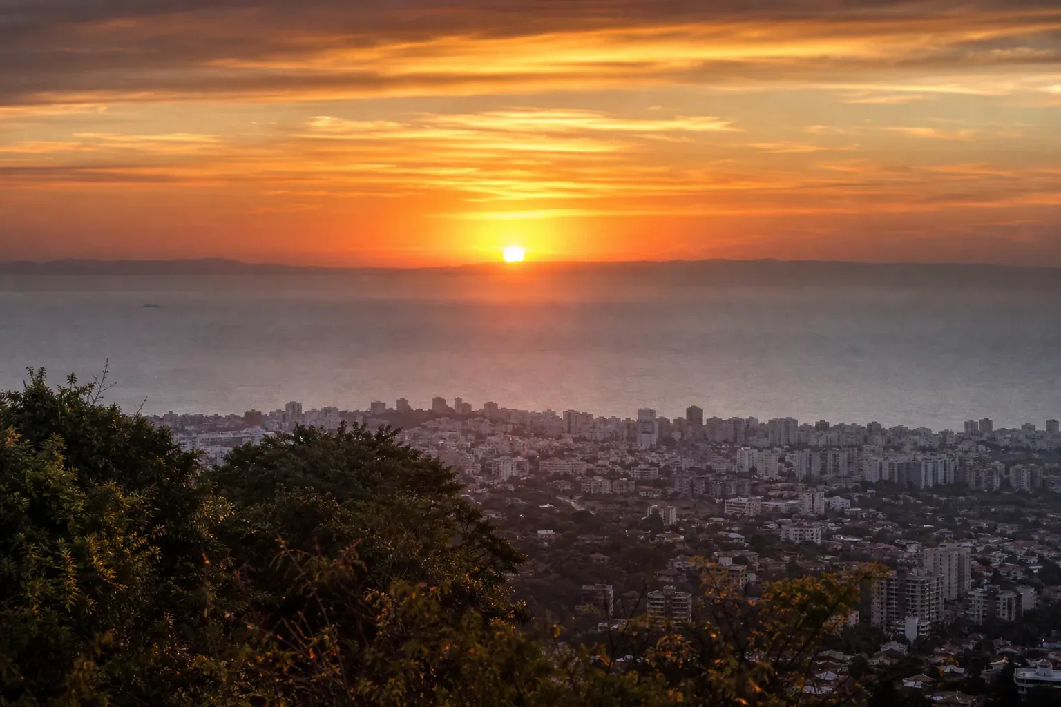 Un coucher de soleil orange intense au-dessus d’une ville côtière, avec des nuages fins et une légère brume atmosphérique.