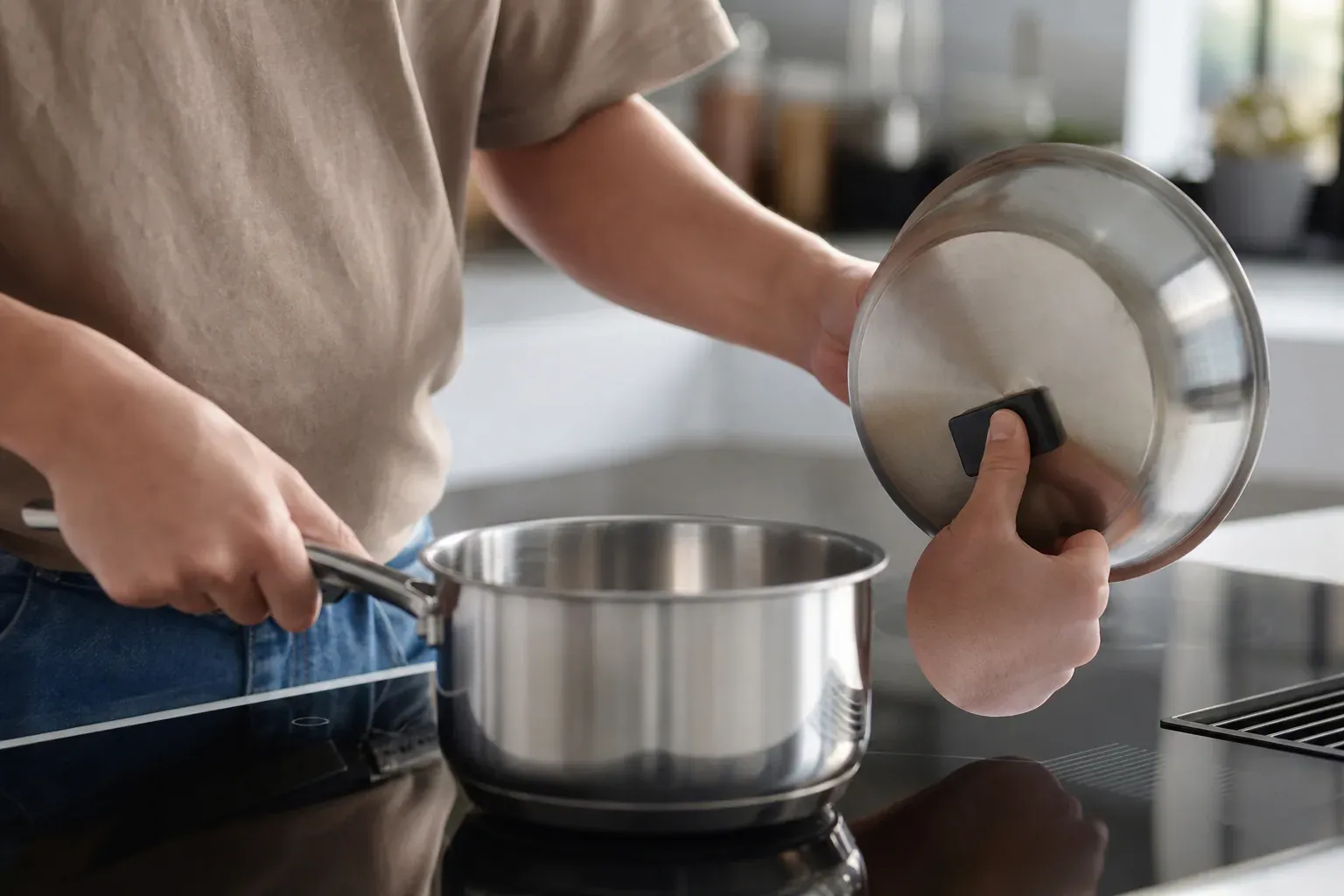 Personne posant une casserole en inox sur une plaque à induction noire dans une cuisine moderne, couvercle à la main.