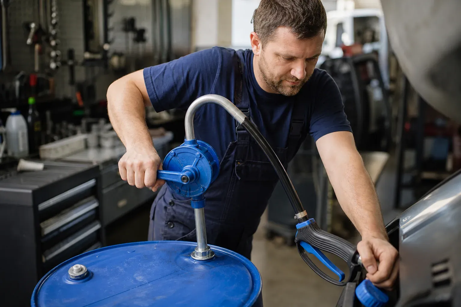 Un technicien de garage transfère de l’AdBlue depuis un fût bleu avec une pompe rotative manuelle dans un atelier automobile.