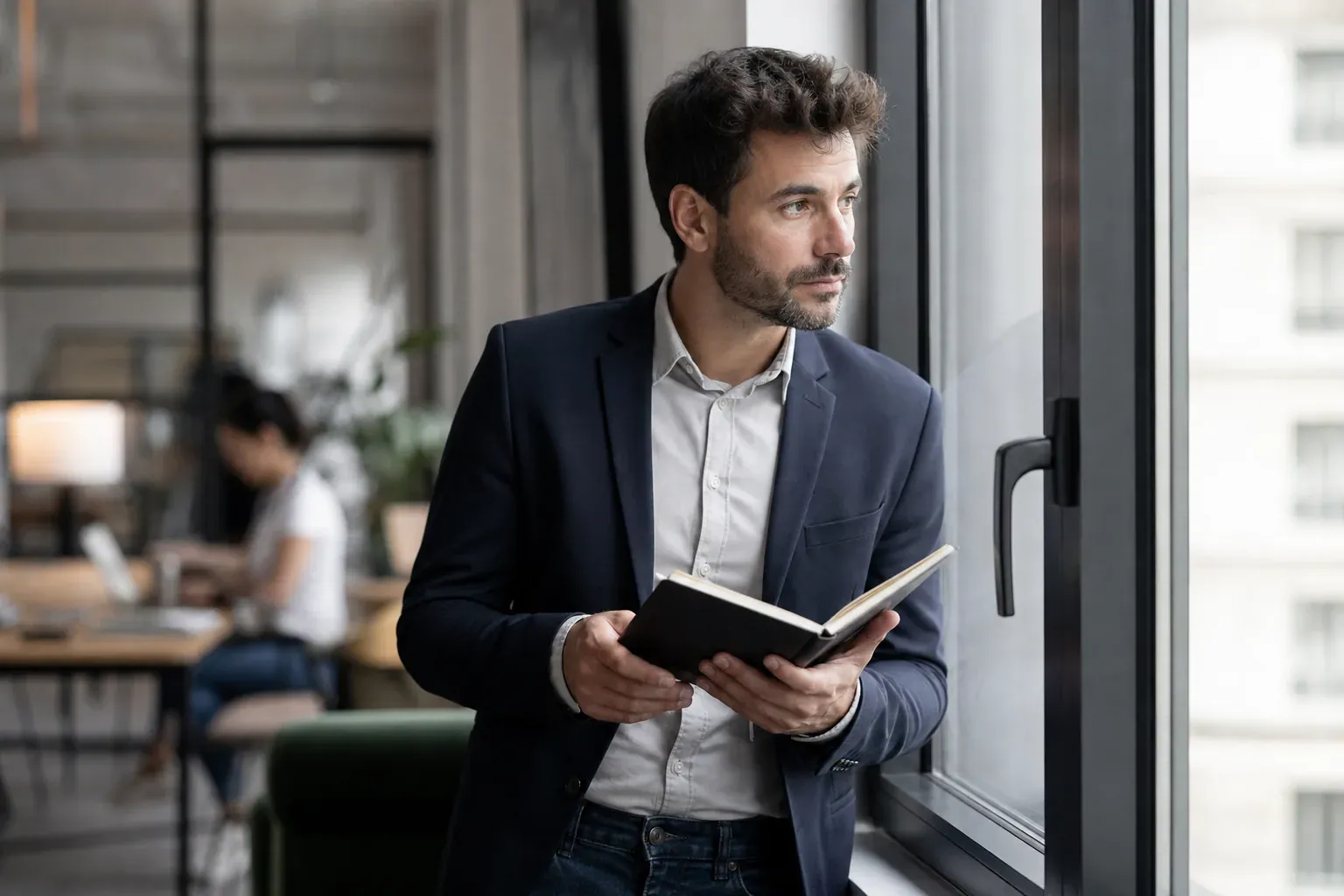 Une personne réfléchit dans un environnement de travail moderne, carnet en main, devant une grande baie vitrée.