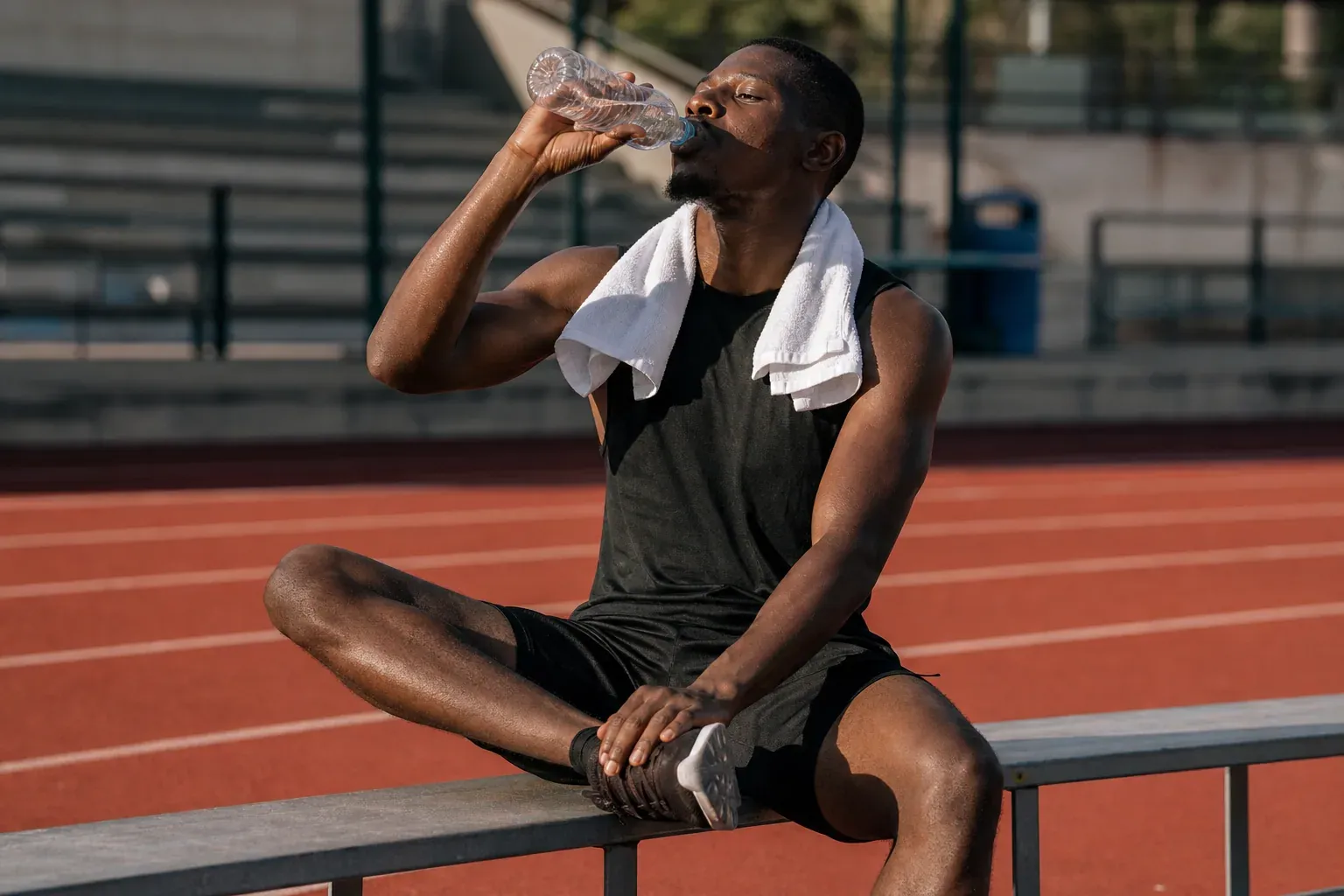 Sportif en phase de retour au calme après l’effort, assis sur un banc, en train de boire de l’eau et de s’étirer légèrement.