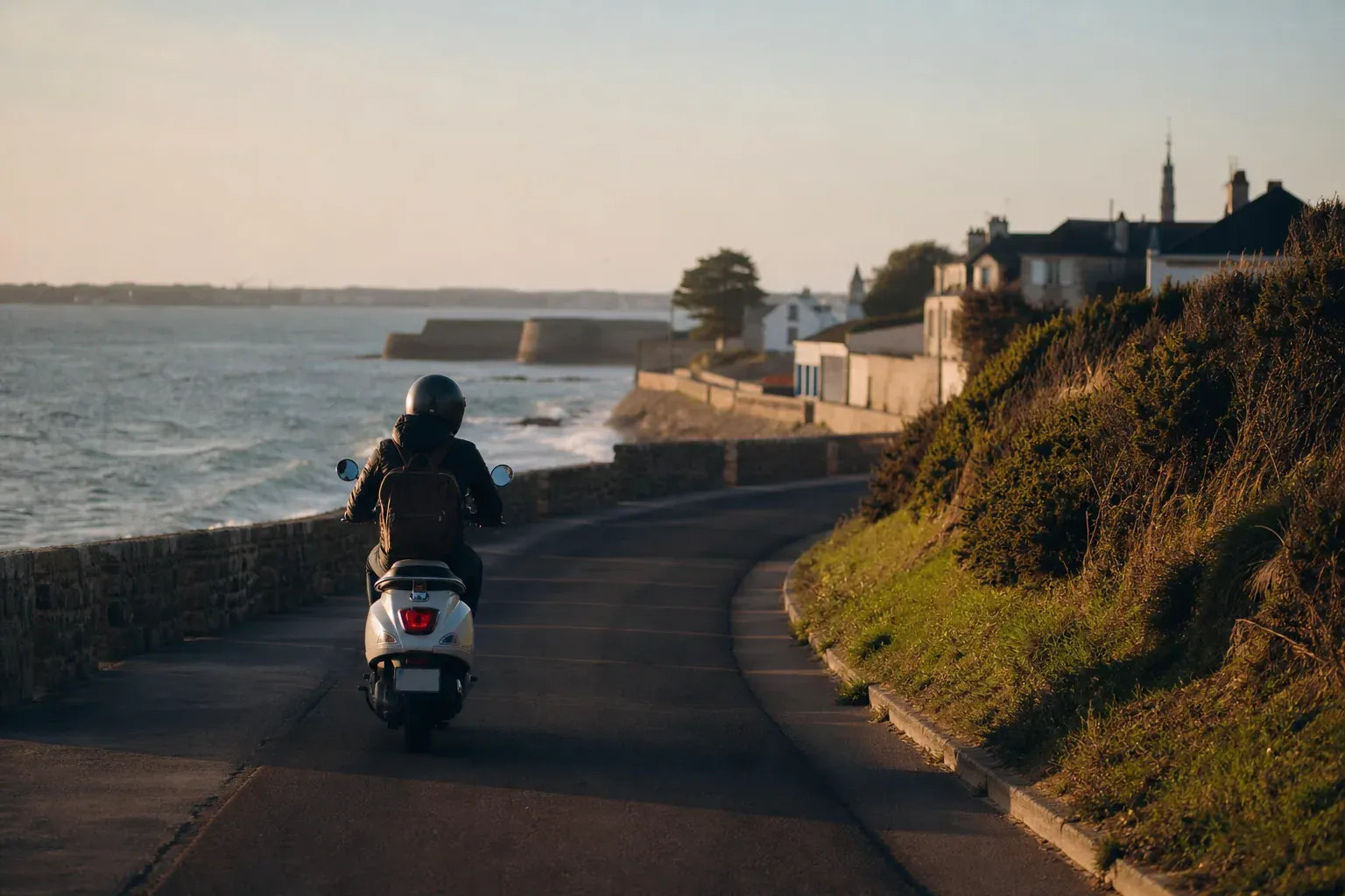 Un scooter roule sur une route côtière près de Port-Louis, avec la rade de Lorient et la mer en arrière-plan au coucher du soleil.