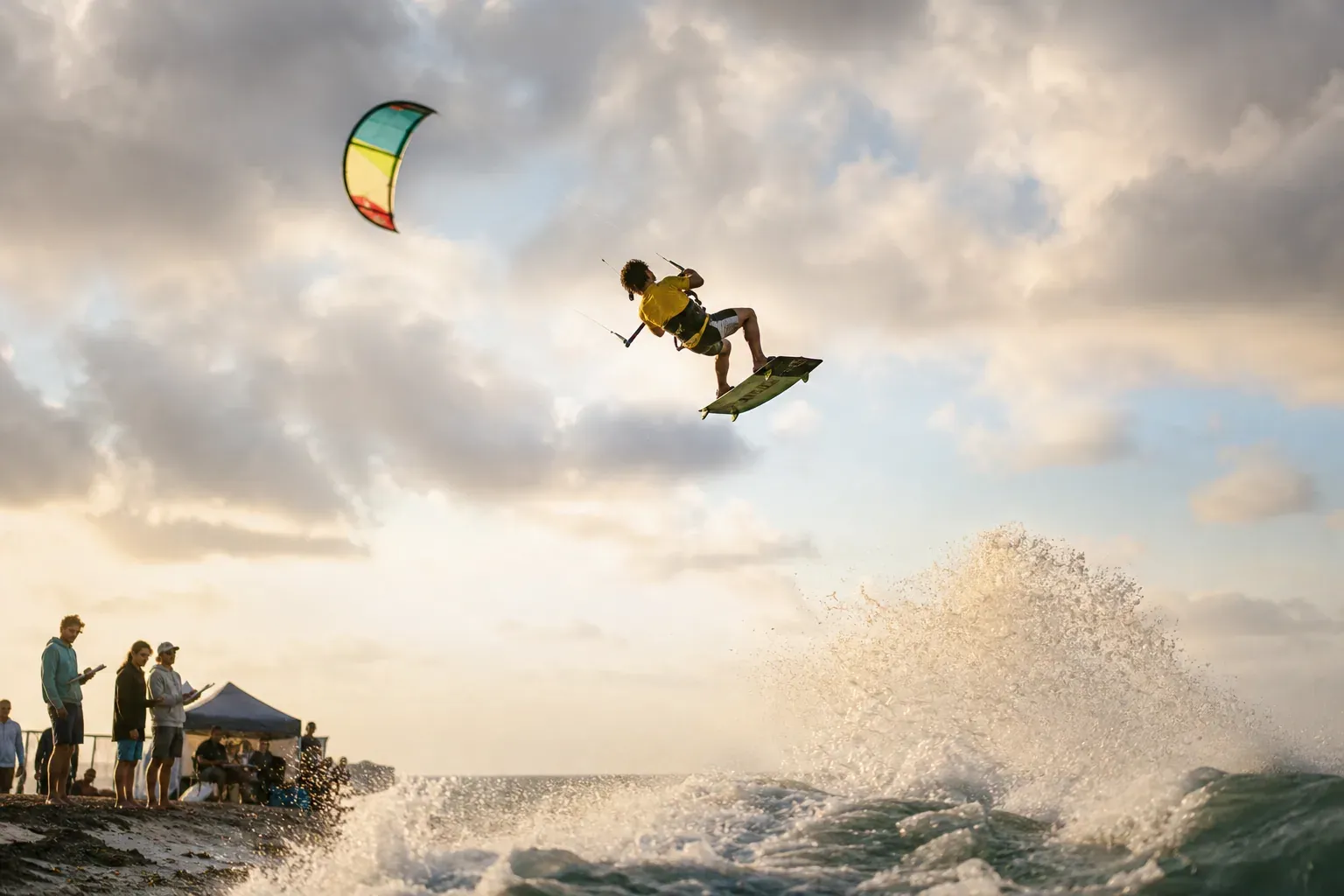 Un rider de kitesurf en plein saut au-dessus de l’eau pendant une compétition, avec le vent, les vagues et des spectateurs en arrière-plan.